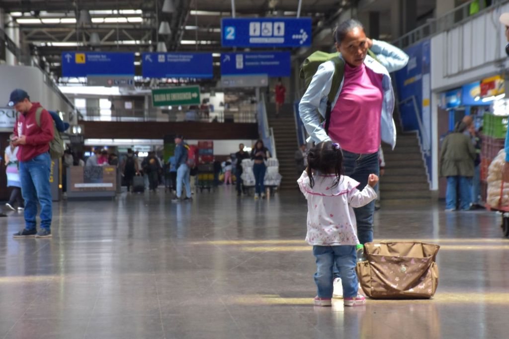 Una madre con una niña pequeña en un terminal de transporte, rodeadas de otros pasajeros y señales informativas.
